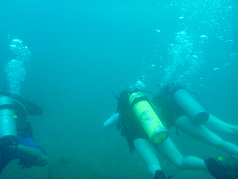 The image shows three scuba divers underwater. The divers are equipped with scuba gear, including tanks and masks. Bubbles are rising to the surface, indicating they are breathing underwater. The water is blue, and visibility appears to be somewhat limited. The divers seem to be exploring the underwater environment, possibly observing marine life or underwater formations.